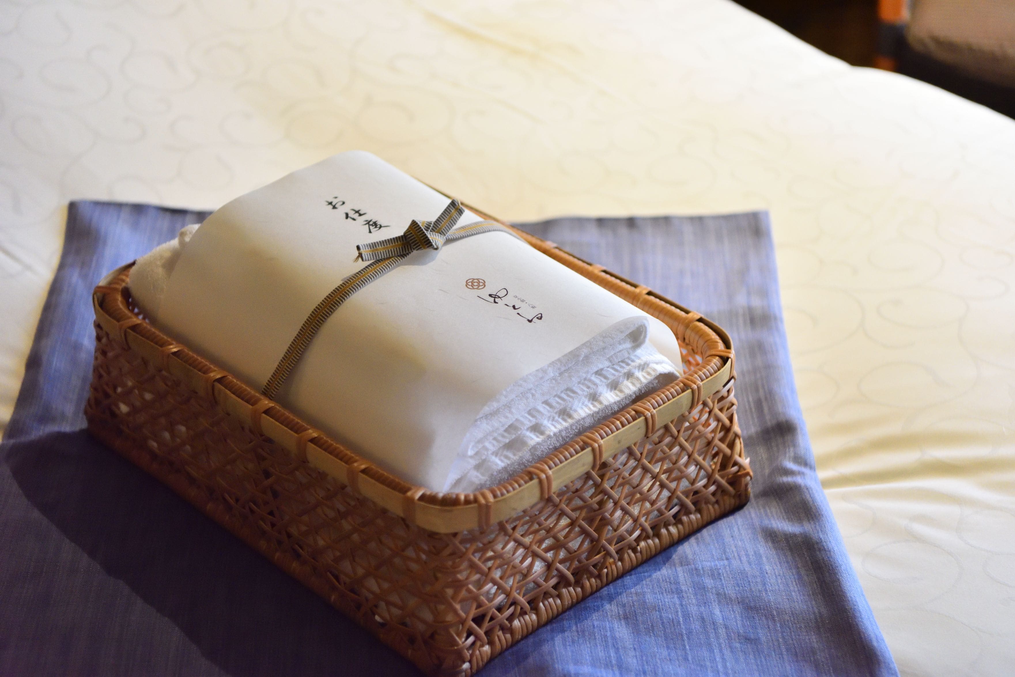 A neatly folded towel with a logo and a ribbon, placed in a woven basket on a bed with a patterned bedspread.