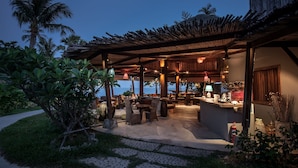 Un patio al aire libre con asientos y bar, rodeado de plantas tropicales y palmeras.