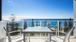 A balcony with a glass table and chairs, overlooking a coastal view with trees and the ocean.