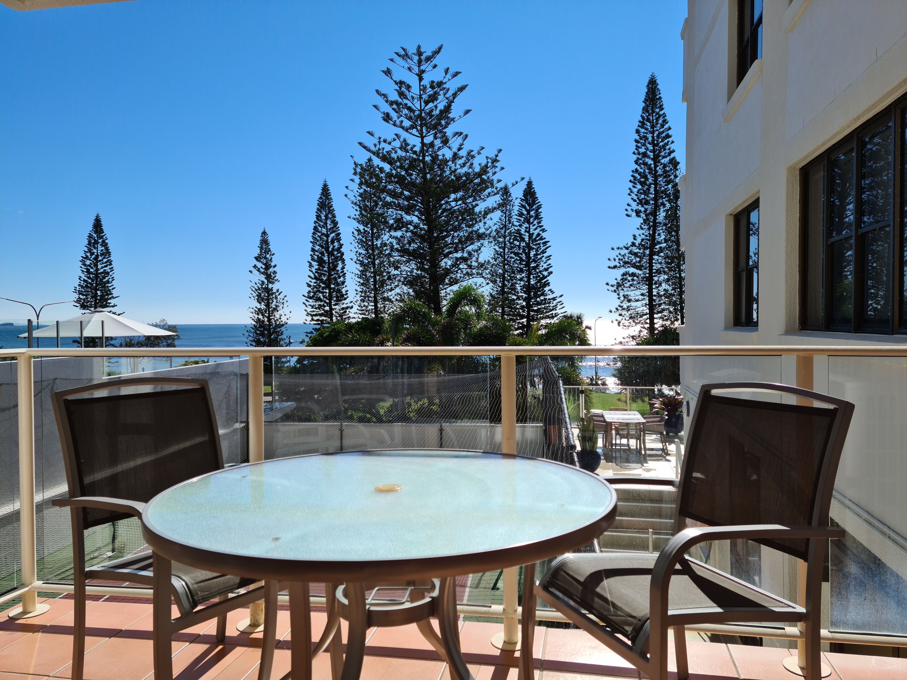 A balcony with a glass table and chairs, overlooking a coastal area with tall trees and a clear blue sky.