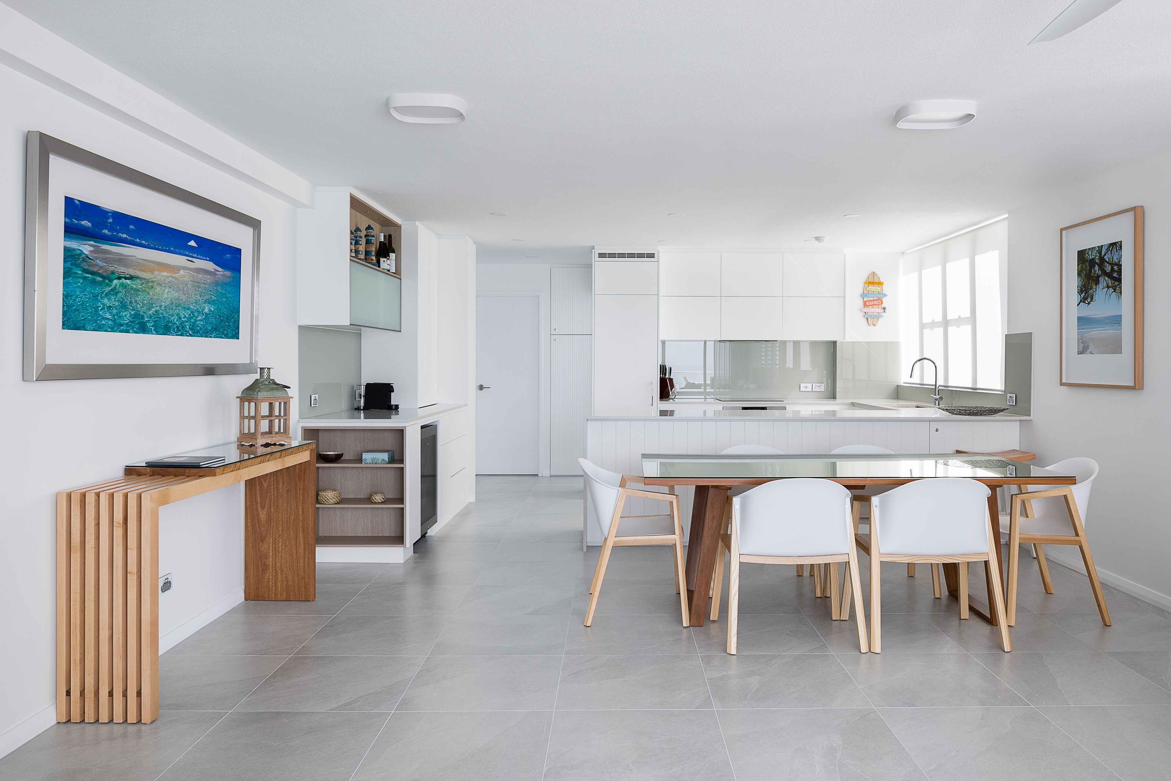 A modern kitchen with white cabinetry, a dining area with a wooden table and white chairs, and a view of the ocean through large windows.
