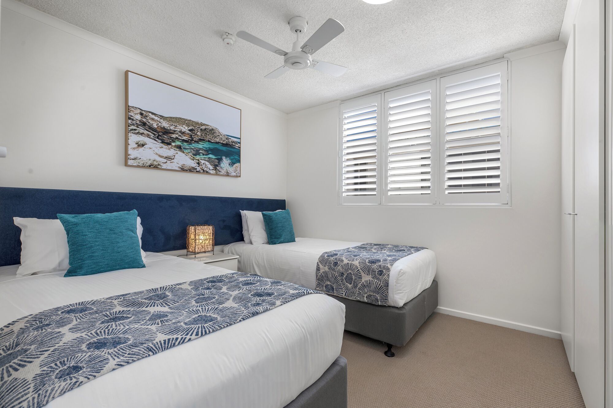 A bedroom with two beds, a ceiling fan, a painting, and a window with blinds.