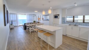 A modern kitchen with a central island, bar stools, and a dining area with a view to the outside. - Pacific Beach Resort (Mooloolaba)