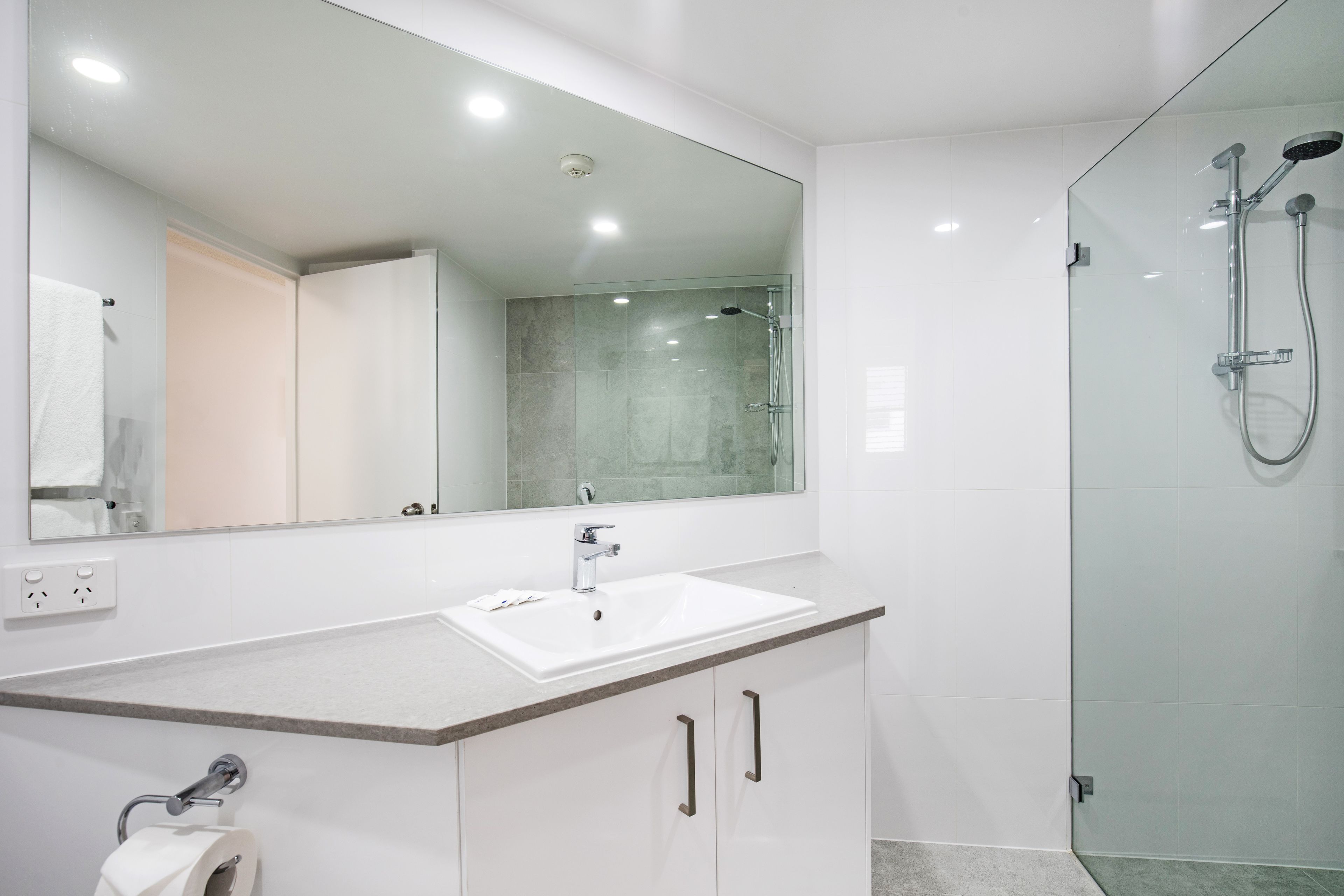 Un baño moderno con un espejo grande, un lavabo blanco y una zona de ducha con mampara de cristal.