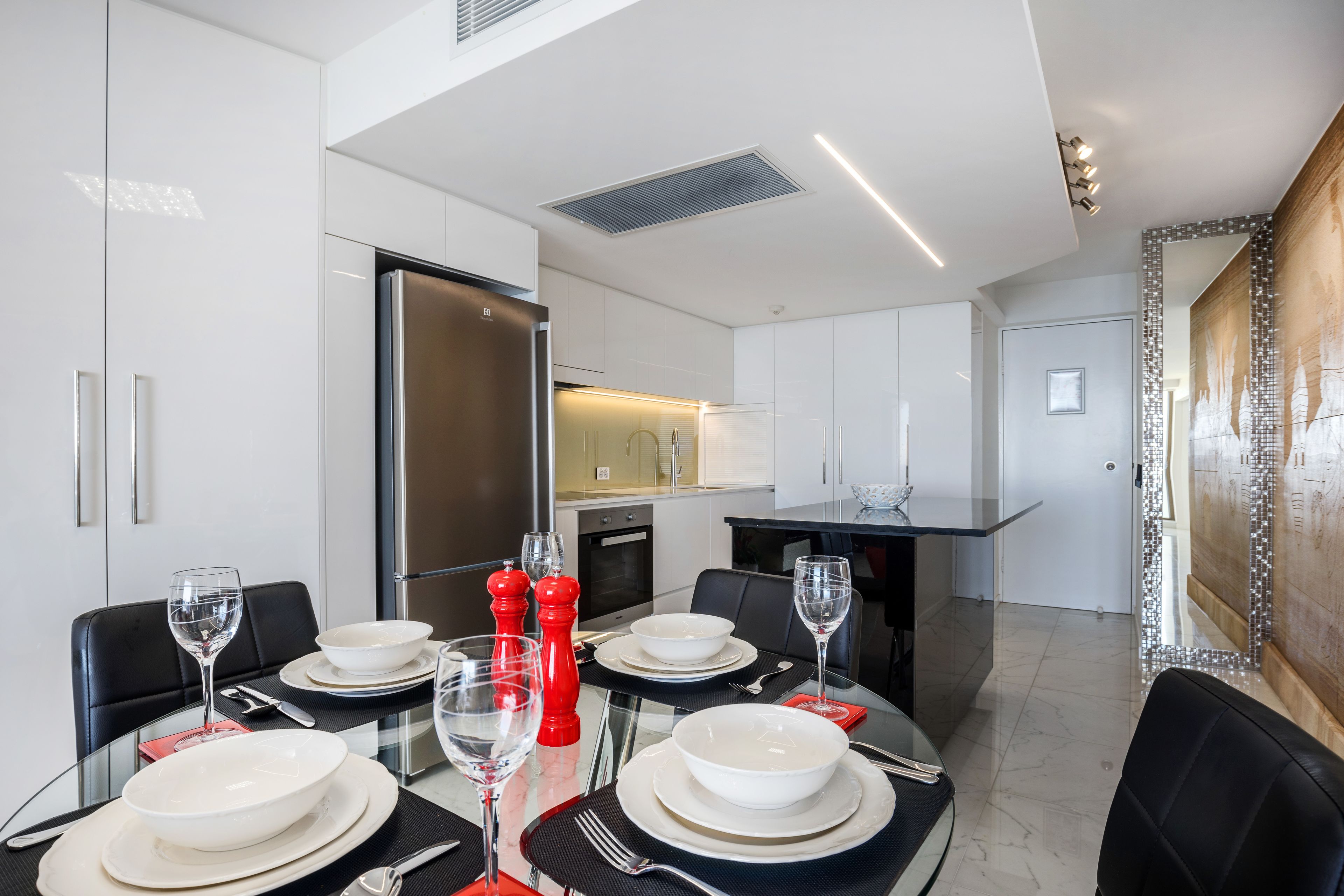 A modern dining area with a glass table set for four, white plates, and red salt shakers.