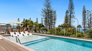 A pool area with lounge chairs and umbrellas, surrounded by tall trees and a clear sky. - Pacific Beach Resort (Mooloolaba)