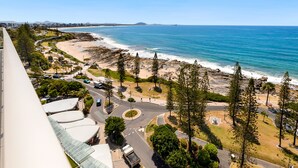 A coastal road with a beach and ocean view, surrounded by greenery and a few buildings. - Pacific Beach Resort (Mooloolaba)