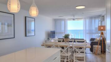 A modern living room with a dining area, large windows, and a view of the sea.