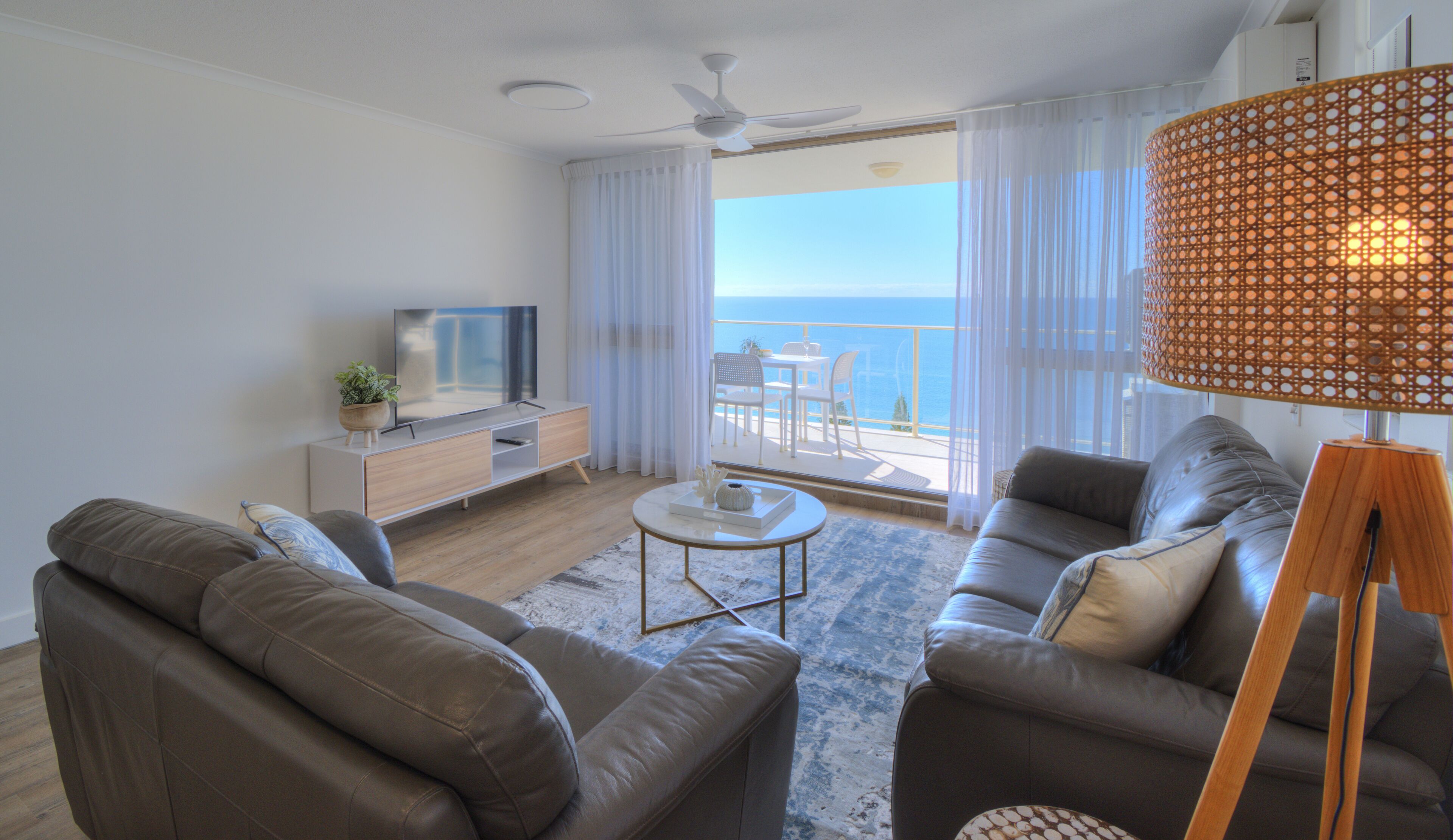 A modern living room with a flat-screen TV, leather sofas, a coffee table, and a large window with a view of the sea.