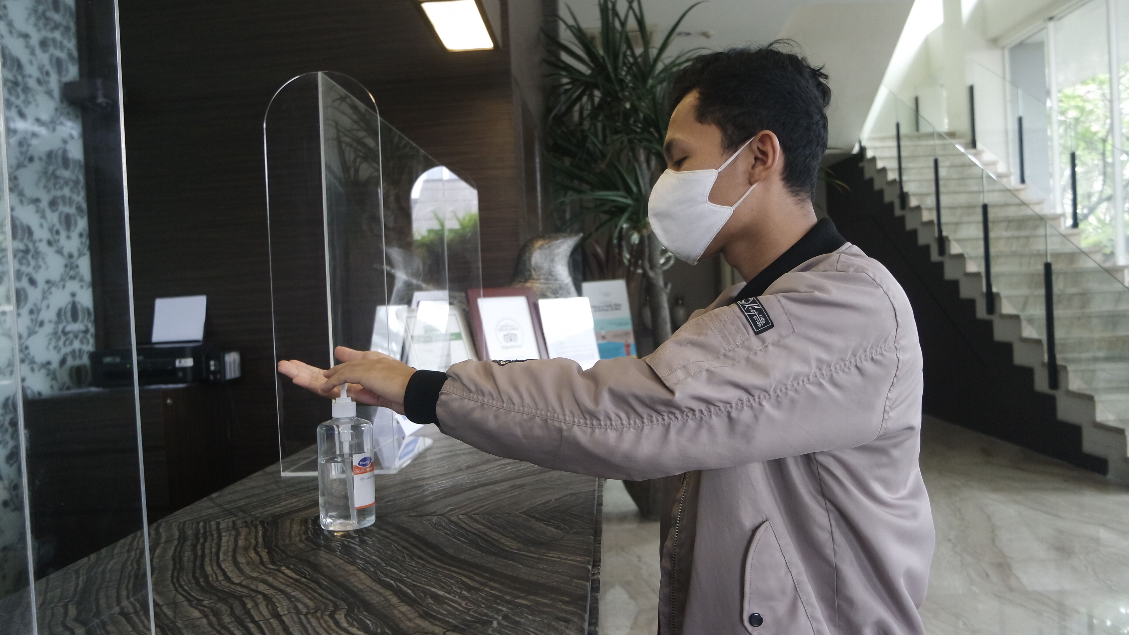 A person using a hand sanitizer dispenser at a reception desk with a modern design, a staircase, and framed certificates in the background.