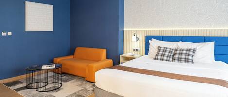 A hotel room with a large bed, a blue accent wall, an orange armchair, a small side table, and a patterned headboard.