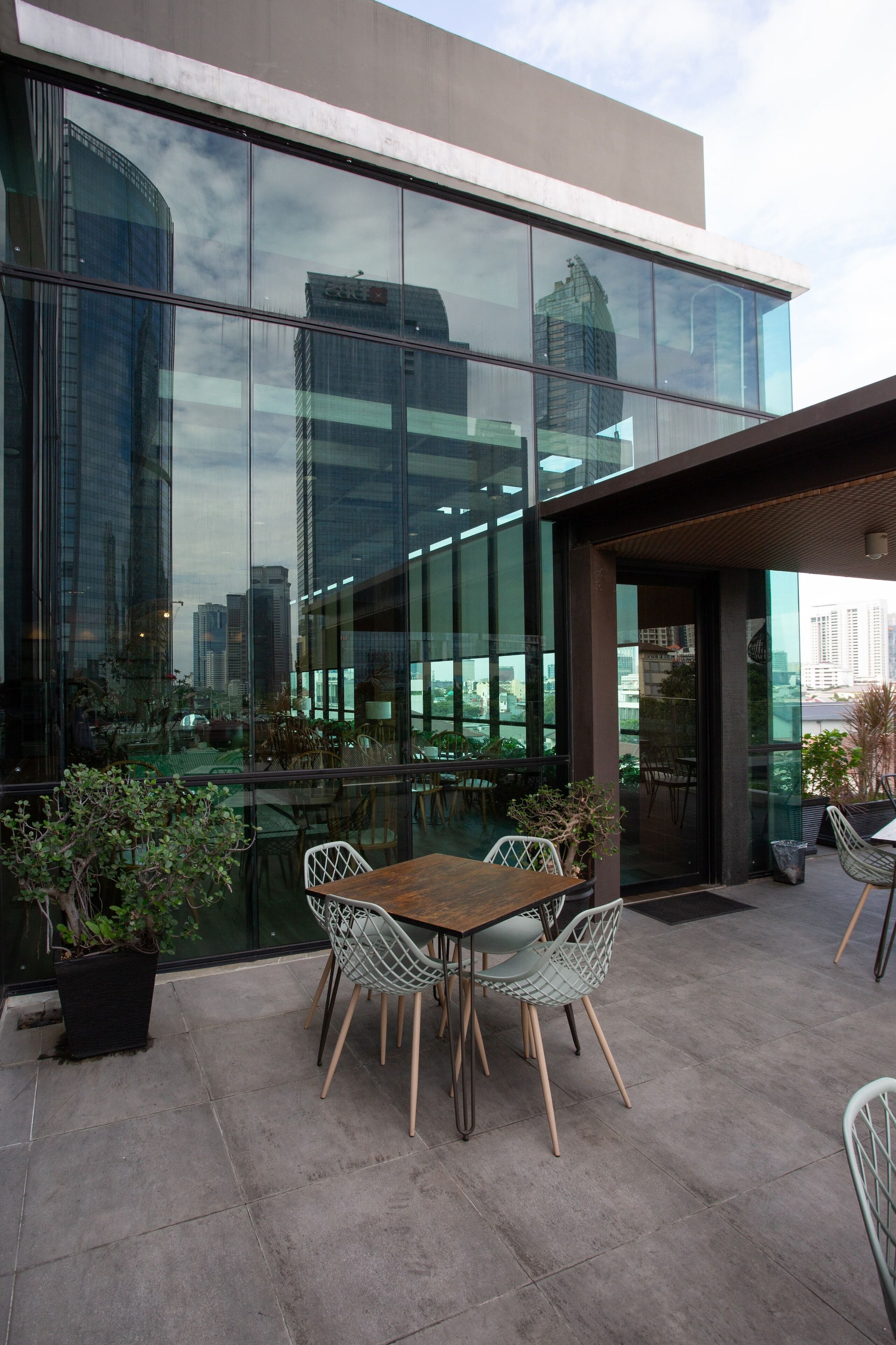 Un área de estar moderna al aire libre con una mesa, sillas, una planta en maceta y vistas de edificios altos a través de grandes ventanales.