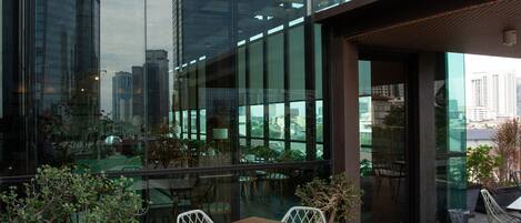 Un área de estar moderna al aire libre con una mesa, sillas, una planta en maceta y vistas de edificios altos a través de grandes ventanales.