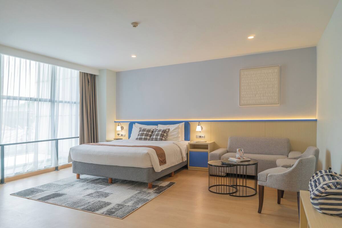 Una habitación de hotel moderna con una cama grande, un sofá, una mesita y vistas a la ciudad a través de amplios ventanales.