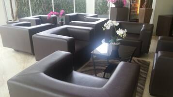 Un área de estar moderna con varios sofás y sillones oscuros dispuestos alrededor de una mesa central. Se observan plantas en macetas y un refrigerador al fondo.