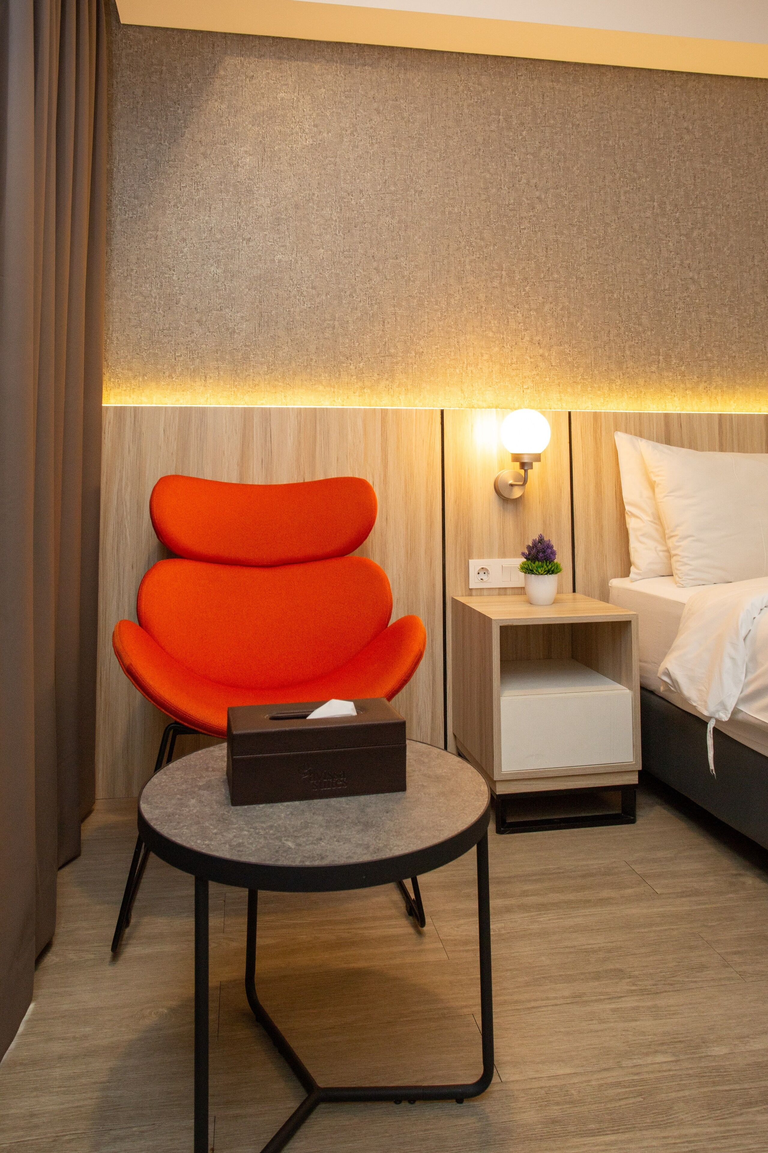 Una habitación de hotel moderna con una pared de madera, una mesa redonda con una caja, una silla naranja, una mesita de noche con una lámpara y una cama con ropa blanca.