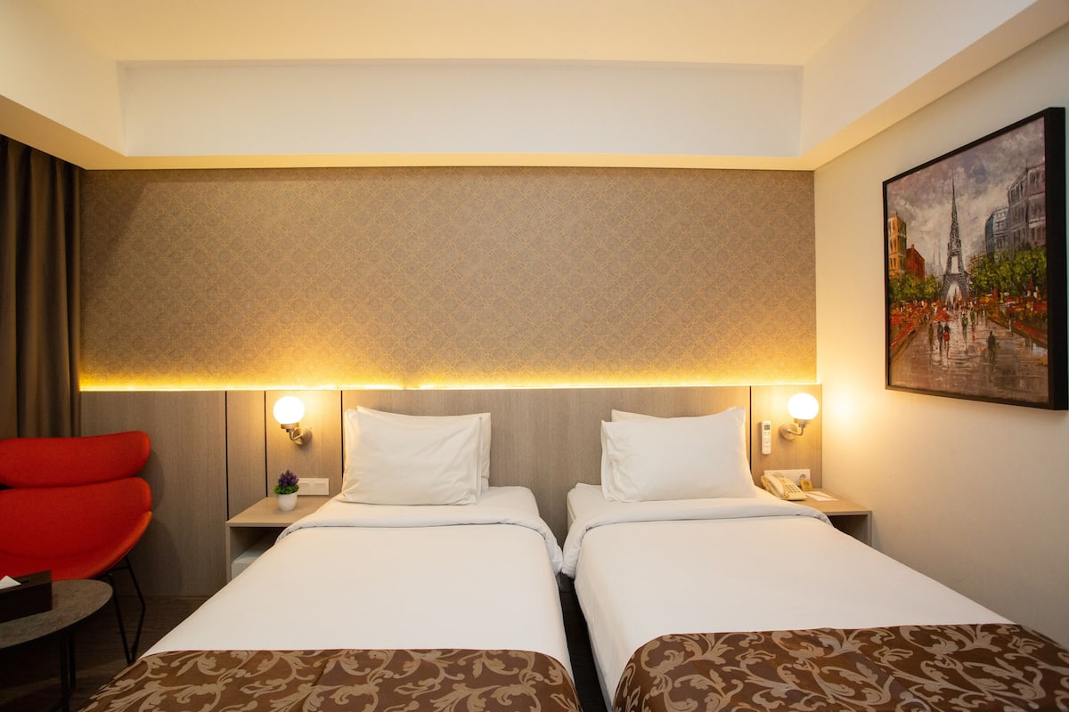 Habitación de hotel con dos camas, una silla roja, una mesita y un cuadro en la pared.