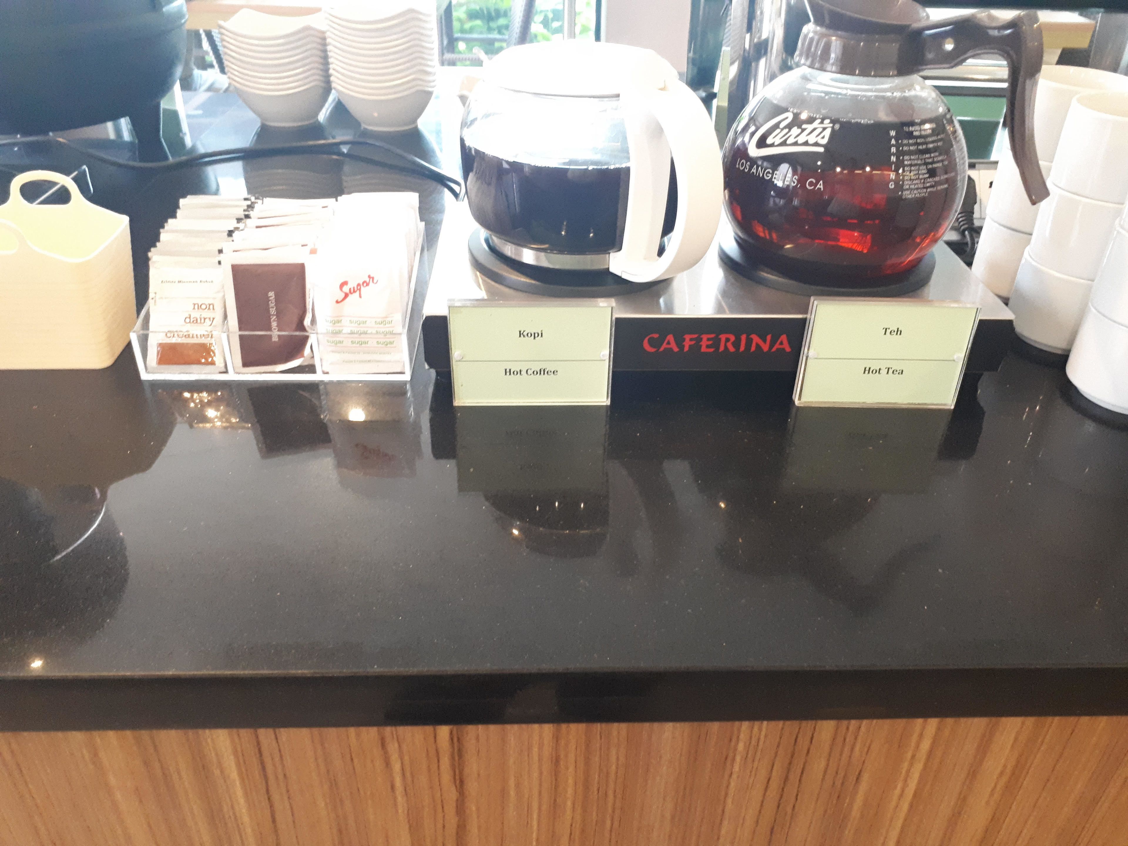 Una estación de café con dispensadores etiquetados para Kopi, Cafarina y Teh, y sobres de azúcar.