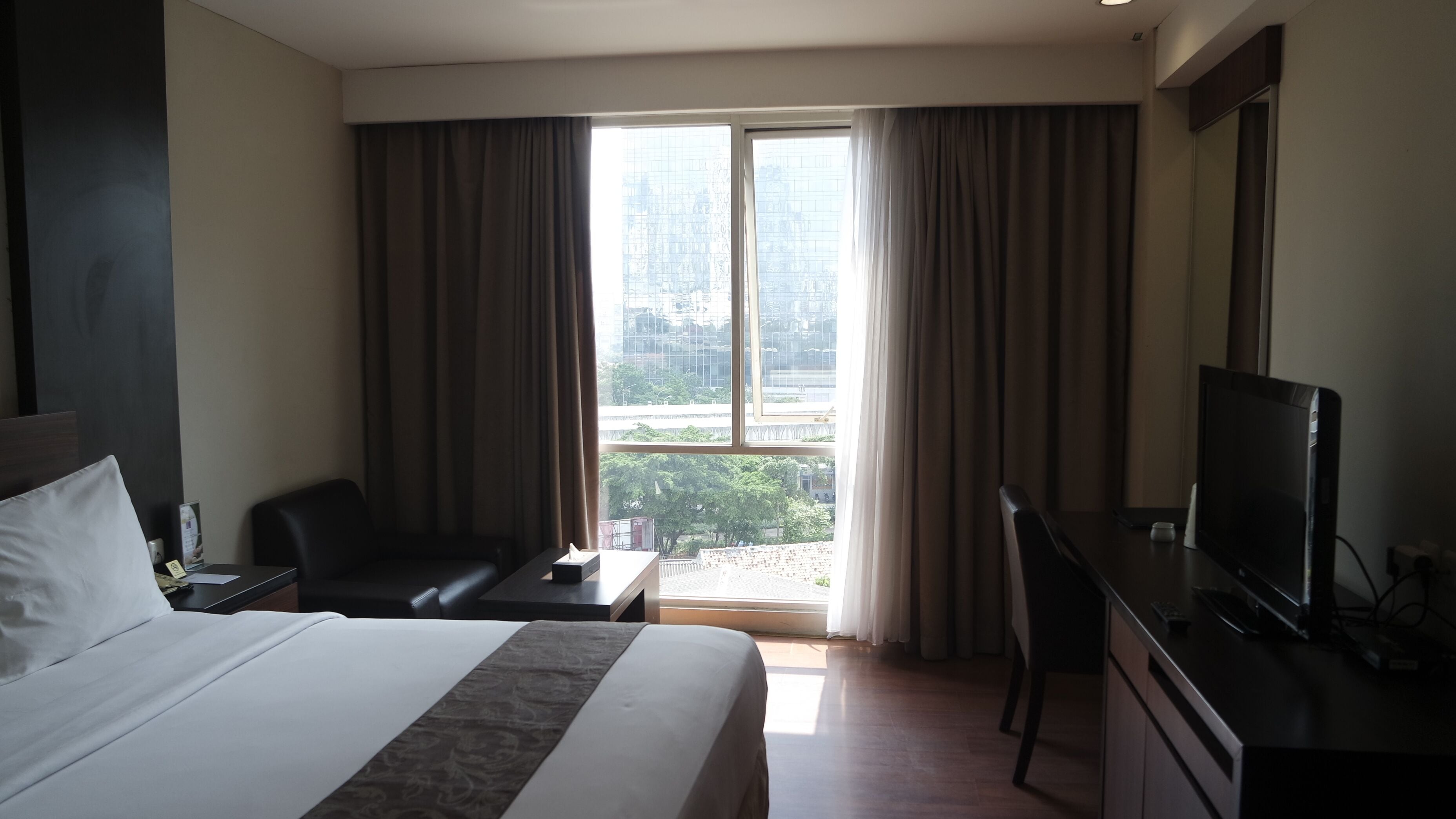 A hotel room with a large bed, a desk with a computer, a TV, and a view of a cityscape through the window.