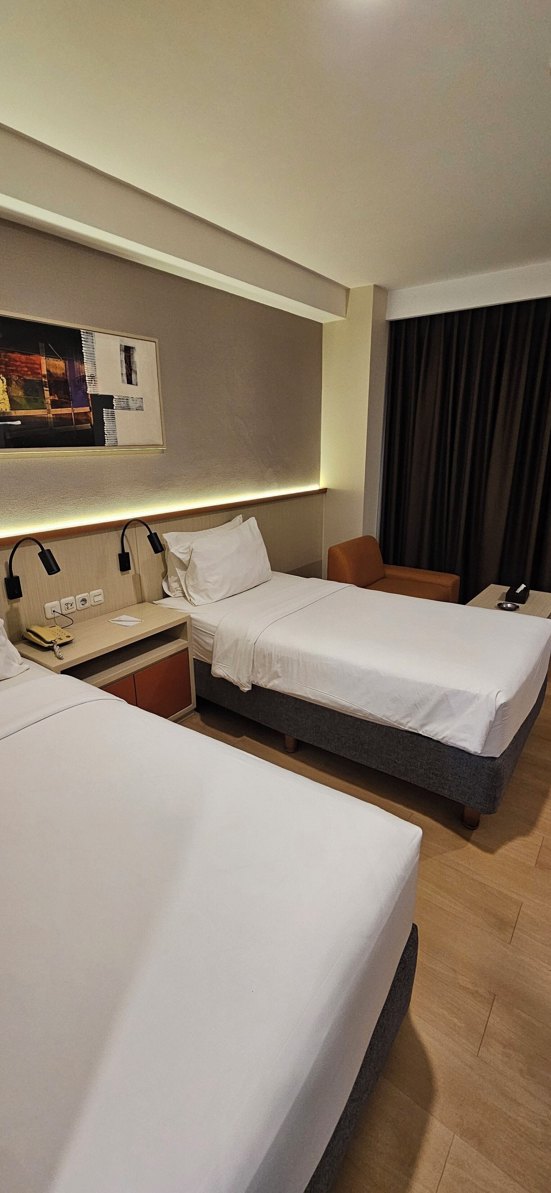 A hotel room with two beds, a bedside table with a lamp, a telephone, and a wall-mounted artwork.