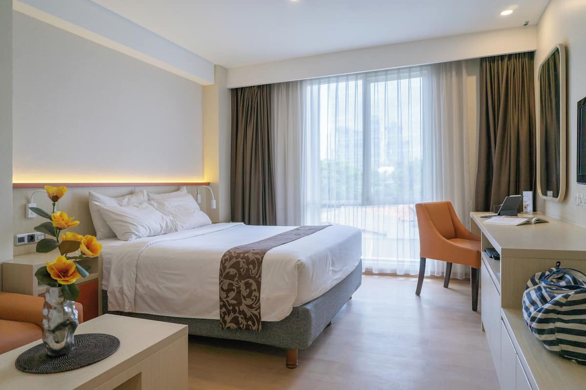 Habitación de hotel moderna con una cama grande, un escritorio con una computadora portátil, una silla naranja y vistas de la ciudad a través de amplias ventanas.