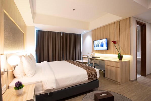 A modern hotel room with a bed, bedside table, lamp, TV, desk, chair, and a small potted plant.
