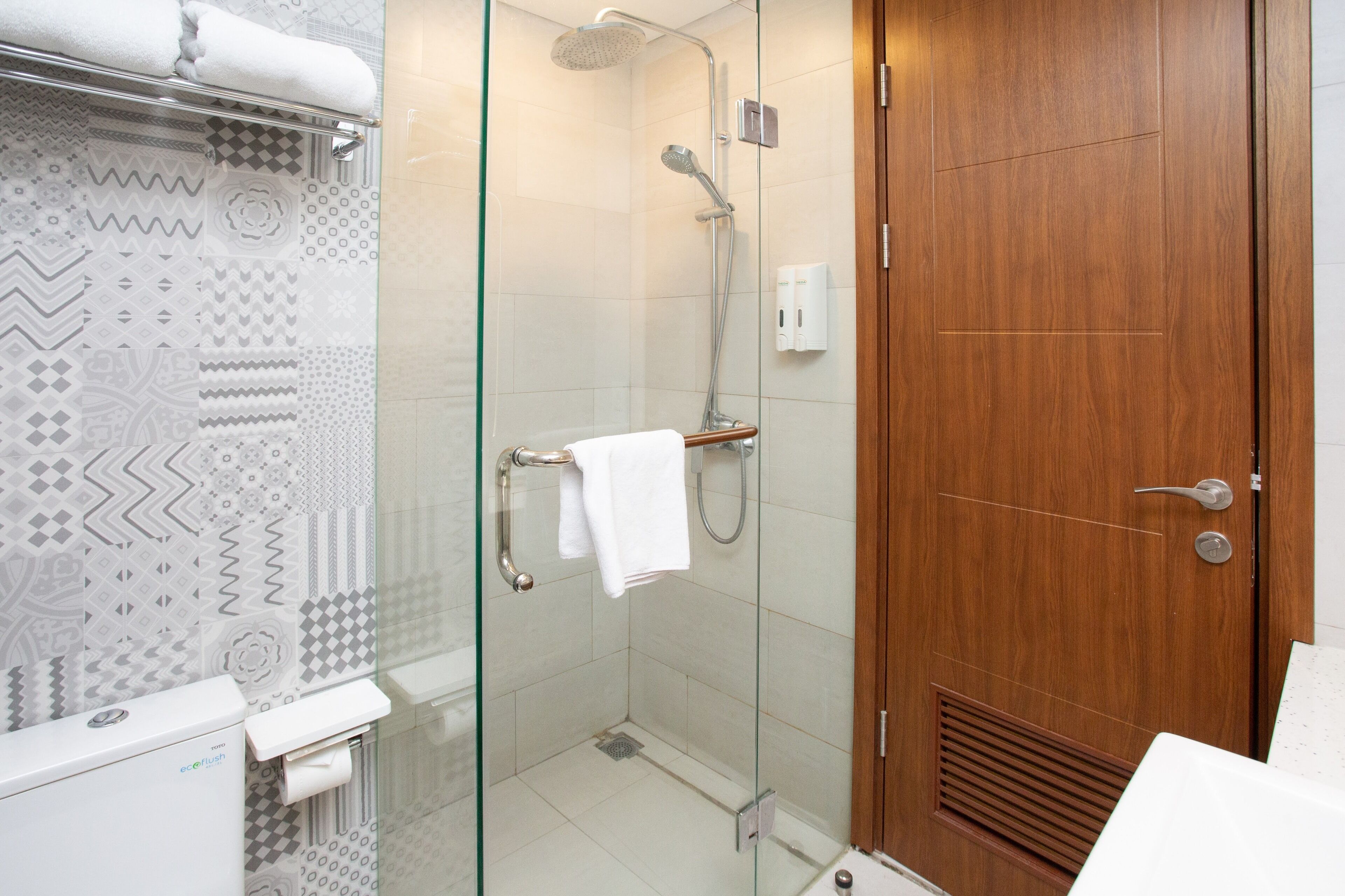 A modern bathroom with a glass shower enclosure, a wooden door, and patterned tiles on the walls.