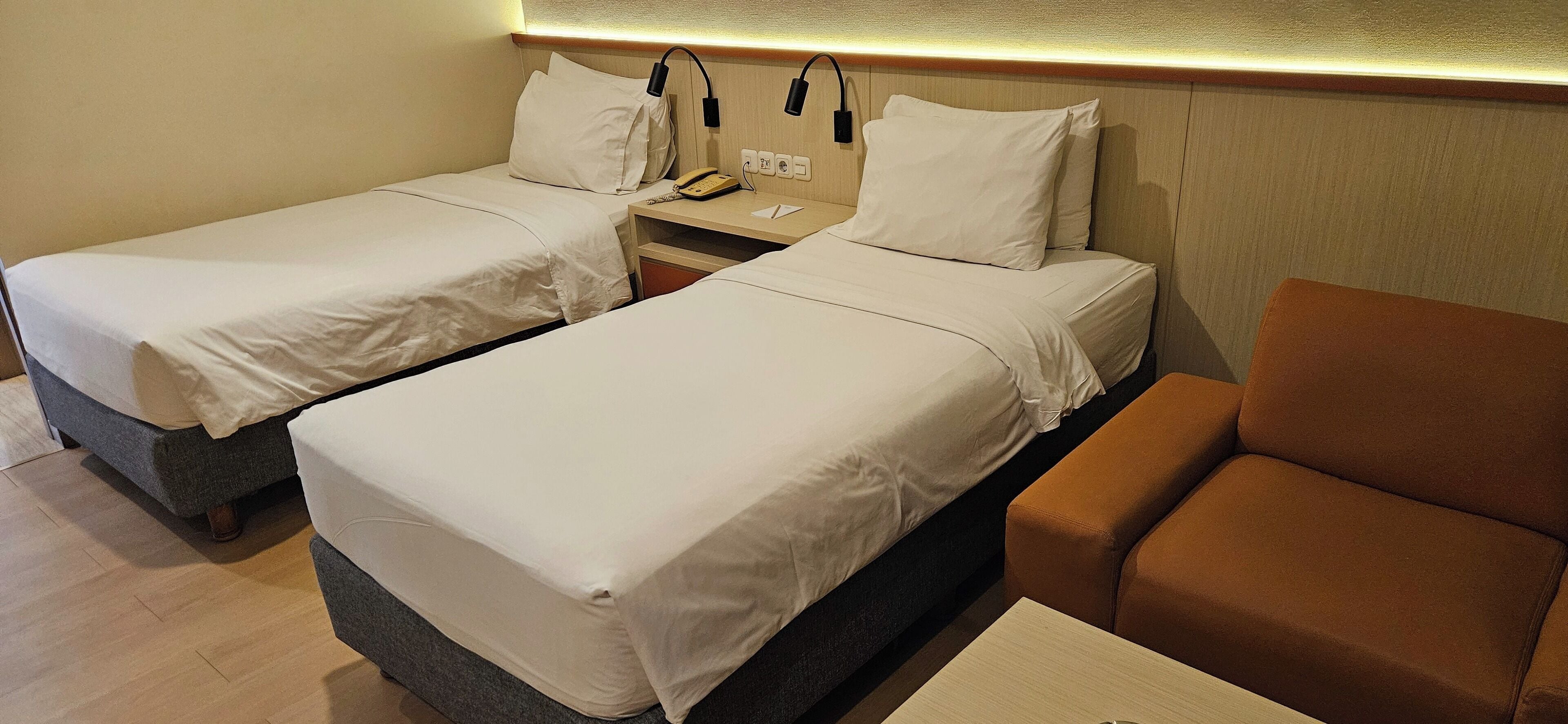 A hotel room with two single beds, a nightstand with a phone and lamp, and a brown armchair.