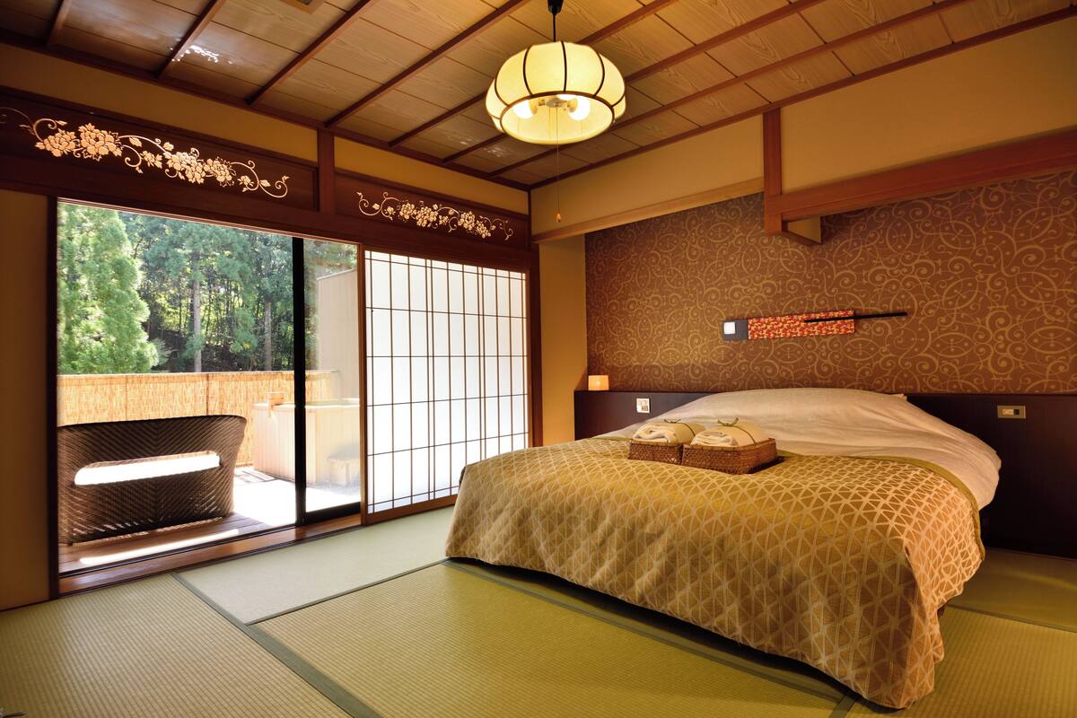 Ein traditioneller japanischer Raum mit Tatami-Boden, einem Bett mit gemustertem Überwurf, einer Schiebetür, die zu einem Balkon führt, und einer Holzbalkendecke mit einer hängenden Leuchte.