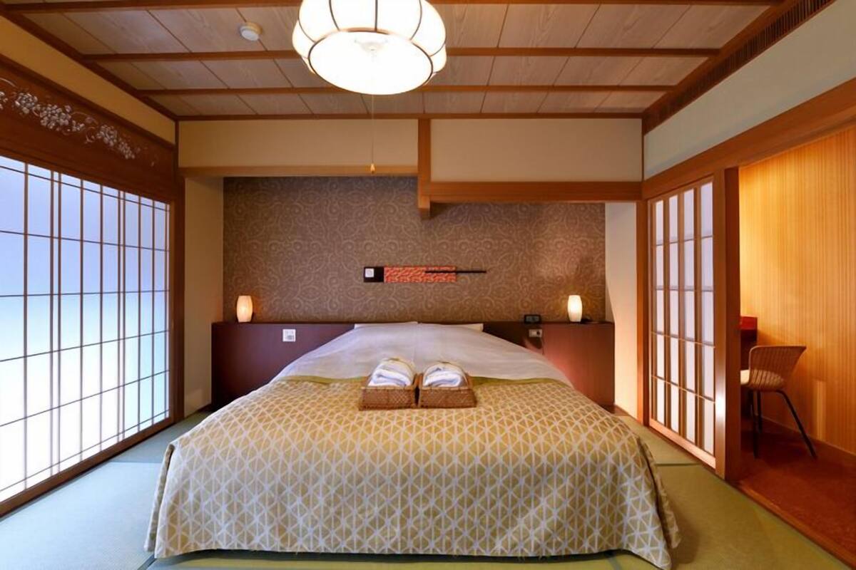 Ein traditioneller japanischer Raum mit Tatami-Böden, einem großen Bett mit gemustertem Überwurf, einem fest montierten Heizkörper und Schiebetüren.