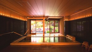 Ein traditionelles japanisches Onsen mit Holzinnenraum, einem Thermalbad und Blick auf einen Felsengarten durch große Schiebetüren.
