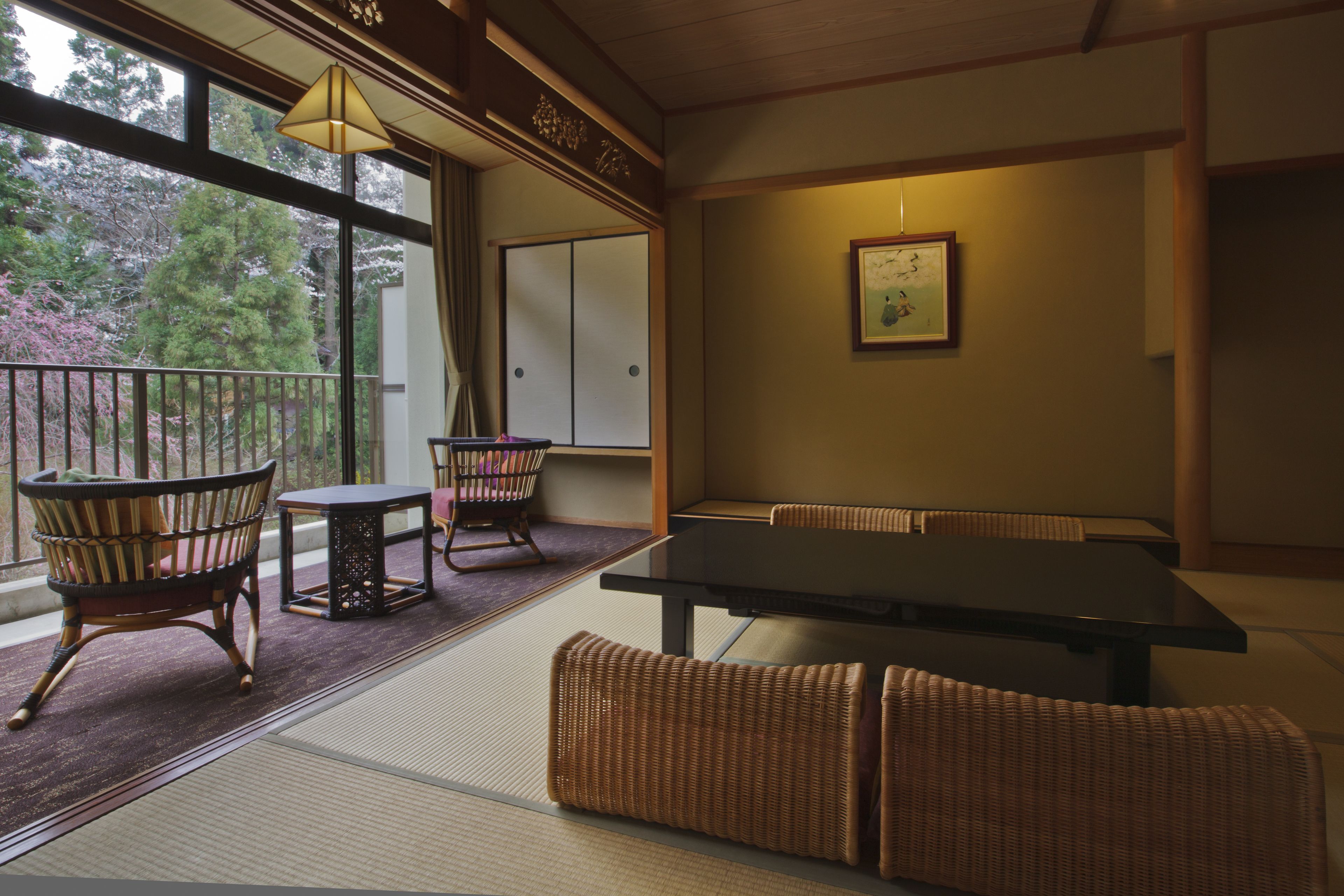 A traditional Japanese room with tatami flooring, a low table, two chairs, and a view of trees through large windows.