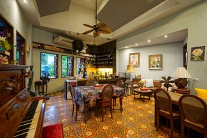 Un salon chaleureux avec un piano, une table à manger et des chaises, agrémenté d’un sol à motifs colorés et d’un ventilateur de plafond.