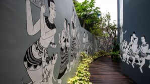 Un pasillo con un mural que representa figuras tradicionales, suelo de madera y una fila de plantas a lo largo de un lado.