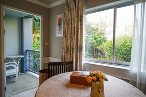 Une pièce avec une table ronde, un vase de fleurs, une boîte, une fenêtre donnant sur un paysage verdoyant et une porte menant à un balcon.