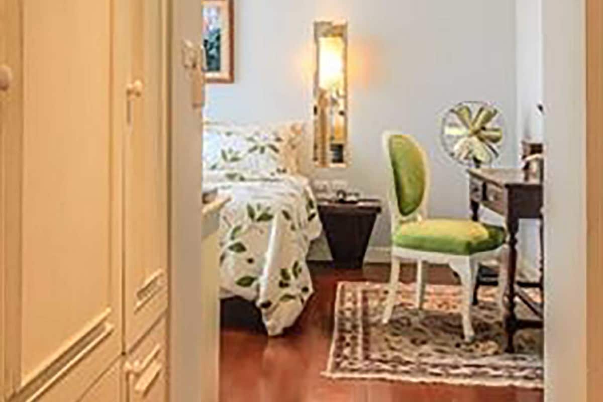 Ein schmaler Flur führt zu einem Schlafzimmer mit Bett, Stuhl, Nachttisch, Lampe und einem gerahmten Bild an der Wand.