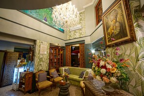 Une pièce avec un canapé vert, des chaises en bois, un lustre, un grand tableau et un arrangement floral.