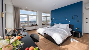 Una habitación de hotel moderna con una cama grande, una mesa de comedor con fruta y flores, vistas de la ciudad a través de amplias ventanas y una pared decorada en azul.