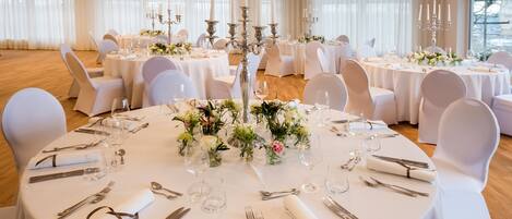 Un salón de banquetes con mesas redondas dispuestas para el servicio de comidas, cada una con mantel blanco, cubertería y cristalería.