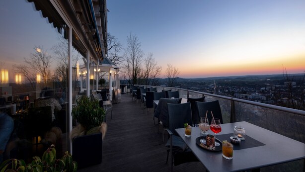 Un área de comedor al aire libre con mesas y sillas, con vistas a un paisaje urbano al atardecer.