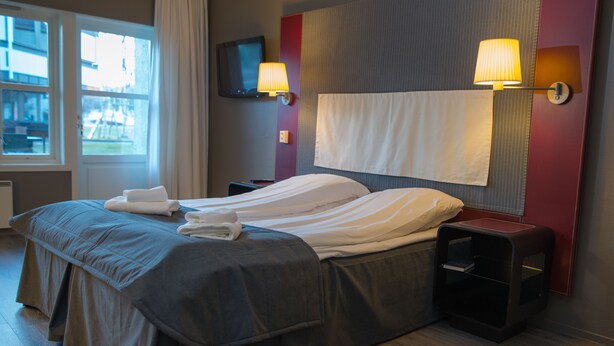 Habitación de hotel moderna con una cama grande, dos mesitas de noche, un televisor de pantalla plana y un ventanal con cortinas.