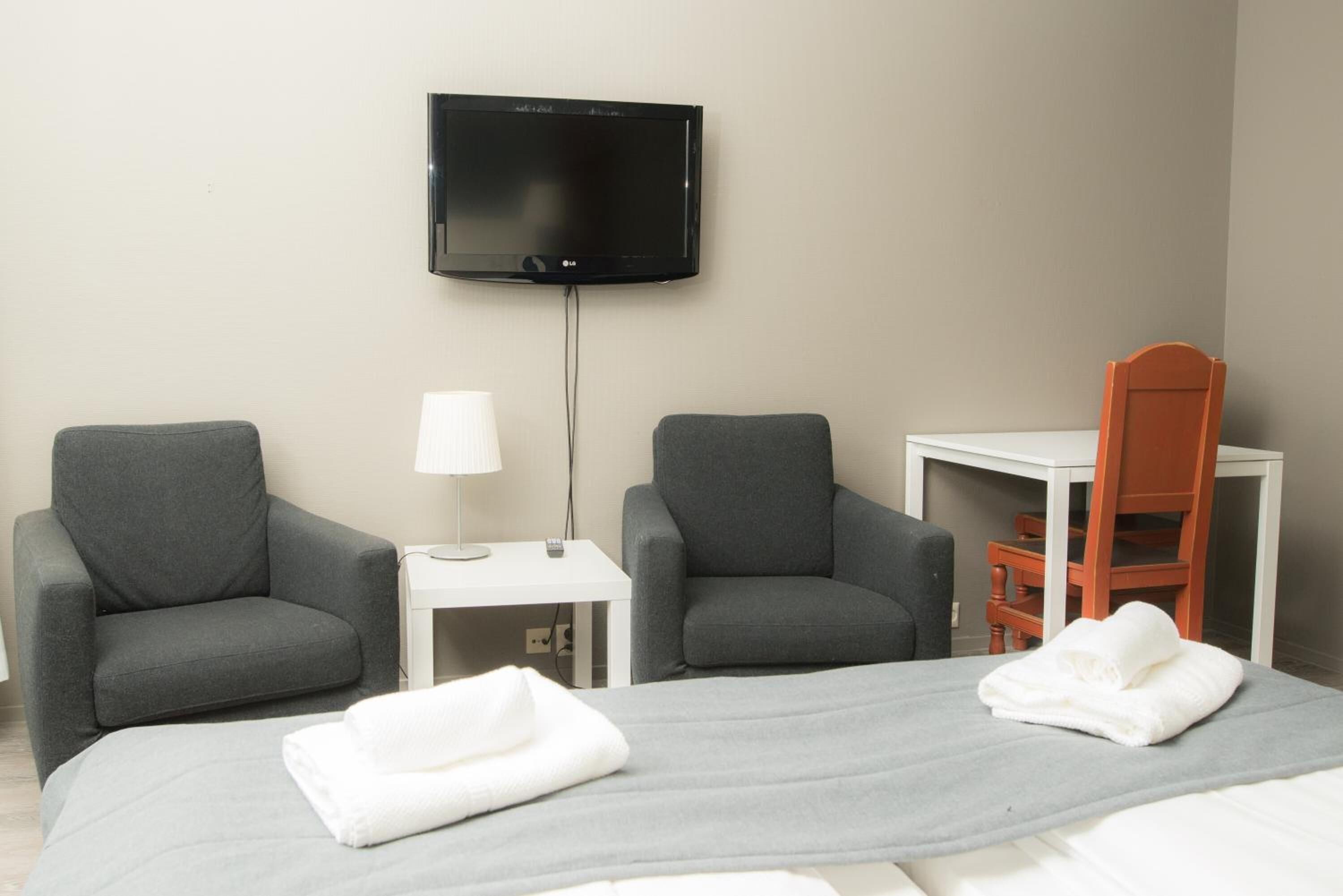A hotel room with two grey armchairs, a small white table with a lamp, a TV mounted on the wall, and a bed with two white towels.