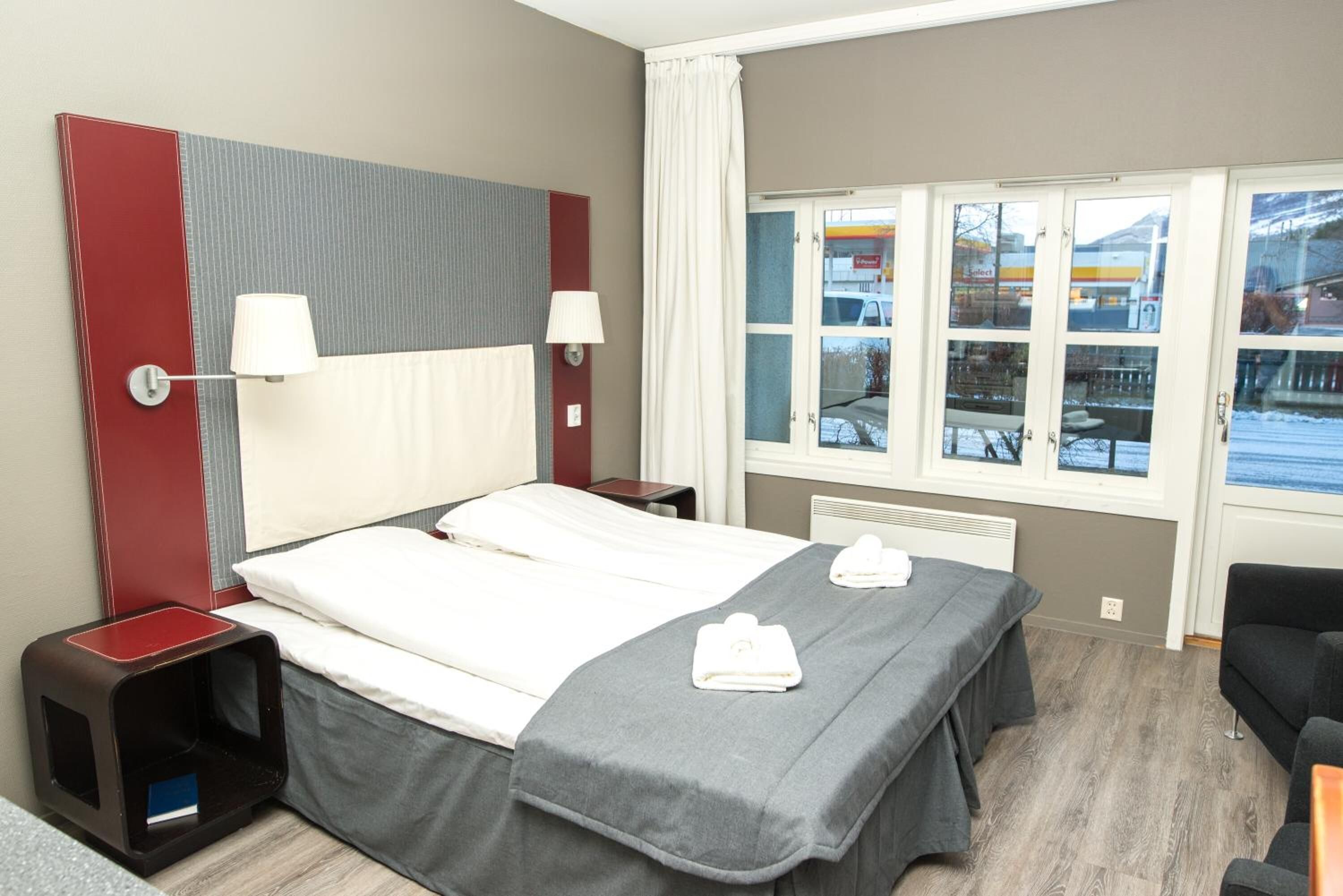 Una habitación de hotel con una cama, mesitas de noche, un ventanal con vistas al exterior y una silla.