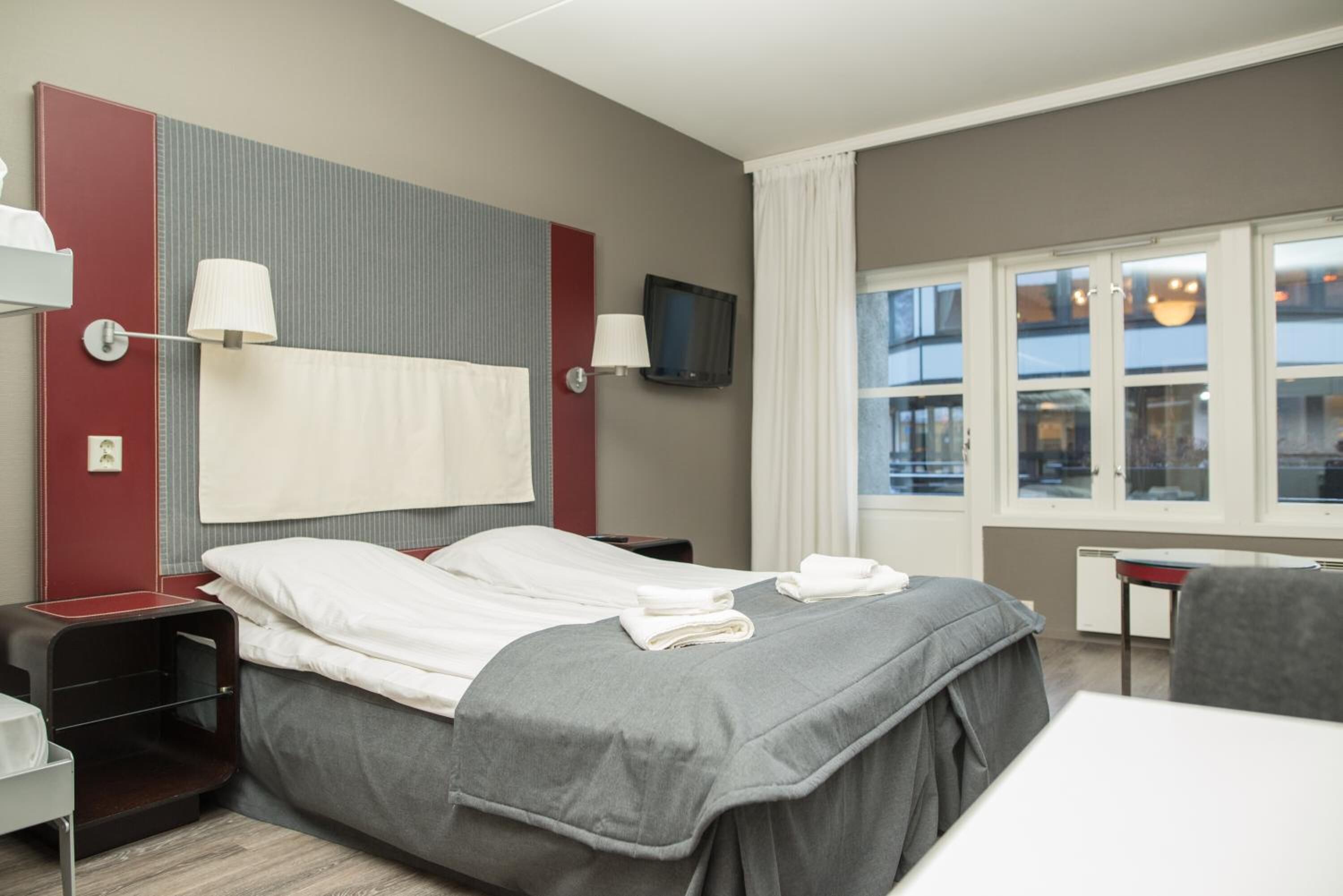 A hotel room with a bed, two bedside tables, a TV, and a window with curtains.