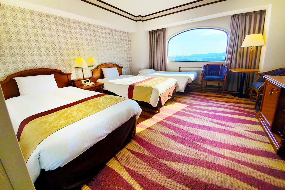 A hotel room with two beds, a desk, a chair, a window with a view, and a patterned carpet.