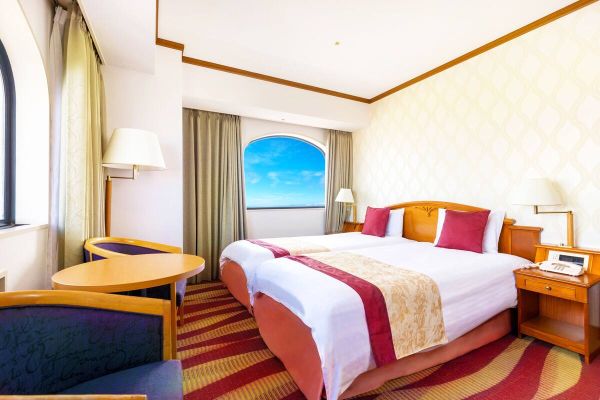 A hotel room with a large bed, a desk, a chair, a nightstand, and a view of the sea through a window.