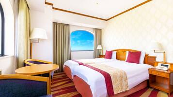 A hotel room with a large bed, a desk, a chair, a nightstand, and a view of the sea through a window.