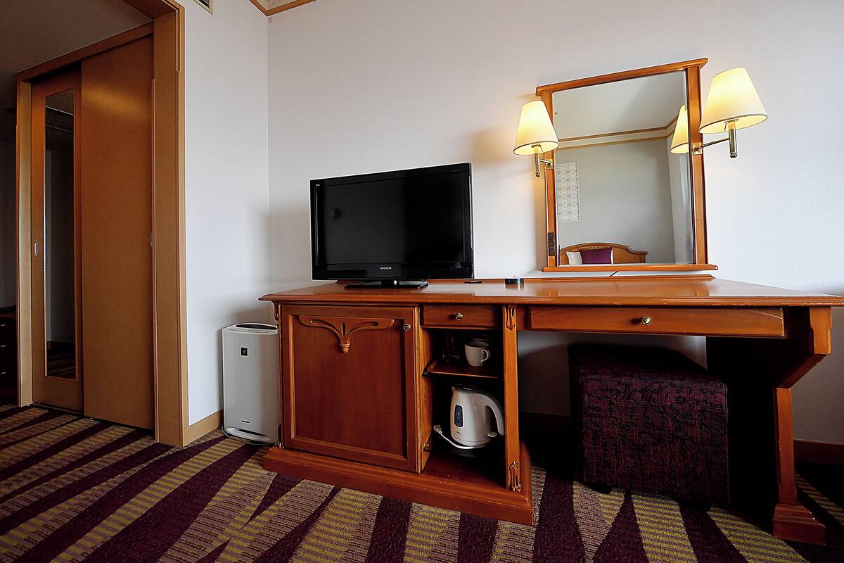 A hotel room with a wooden desk, a television, a mirror, and a lamp.