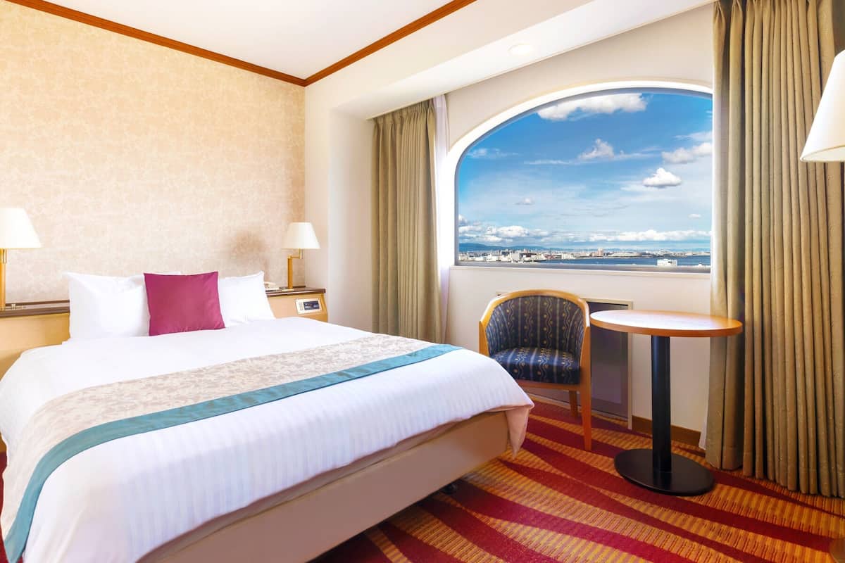A hotel room with a large bed, a chair, a small table, and a view of the city through a large window.