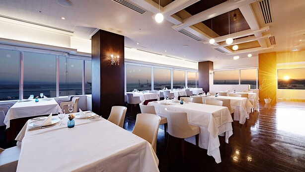Un restaurante con varias mesas, manteles blancos y vistas al atardecer a través de amplias ventanas.